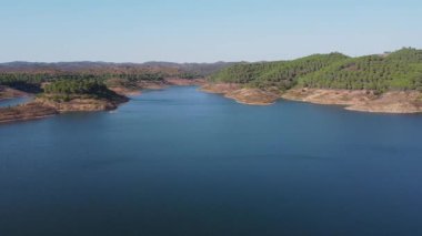Santa Clara barajının berrak mavi suları, çevreleyen tepeleri ve parlak gökyüzünün altındaki yemyeşil yeşil manzarası, Alentejo Portekiz 'deki Santa Clara barajı.