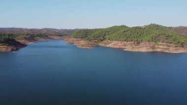 Santa Clara barajının berrak mavi suları, çevreleyen tepeleri ve parlak gökyüzünün altındaki yemyeşil yeşil manzarası, Alentejo Portekiz 'deki Santa Clara barajı.