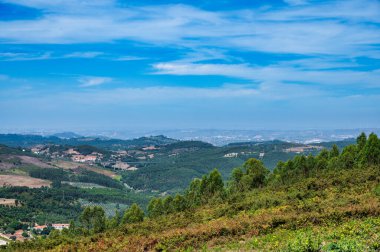 Yuvarlanan tepelerle, yeşil ormanlarla ve bulutlu mavi gökyüzüyle kırsal alan.