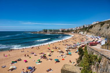 Ericeira, Portekiz. 24 Ağustos 2024. Güneşli plajlarda güneşlenen ve yüzen insanlar, renkli şemsiyeli canlı kıyı manzaraları, berrak gökyüzü, Portekiz 'in Ericeira köyündeki Güney Sahili.