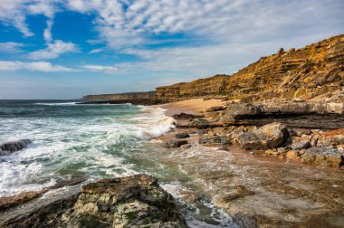 Dalgalar, Portekiz 'in Ericeira kentindeki Pedra Branca' nın kayalık kıyı şeridine katmanlı kayalıklar ve turkuaz su ile çarpar..