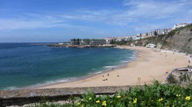 Ericeira 'daki Praia do Sul manzarası, altın kum, turkuaz sular ve canlı bir gökyüzü altında kıyı binaları..