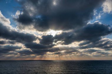 Altın güneş ışığı okyanus üzerindeki kara bulutları delip geçiyor, gün batımında suya ışık saçıyor..