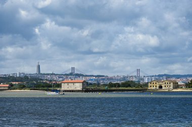 Lizbon 'un Seixal' dan panoramik manzarası, Tejo Nehri ile, dramatik bulutlu gökyüzü altında yelkenli bir tekne, köprü ve anıt..