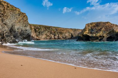 Portekiz 'in Alentejo kentindeki Praia da Samouqueira kumsalında büyük kaya oluşumlarına karşı okyanus dalgaları, parçalı bulutlu bir gökyüzünün altında engebeli bir kıyı manzarasını gösteriyor..