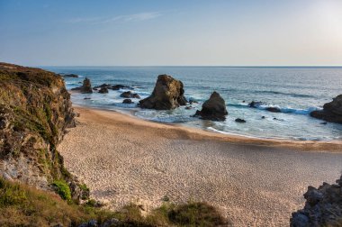 Portekiz 'in Alentejo kentindeki Praia da Samouqueira kumsalında büyük kaya oluşumlarına karşı okyanus dalgaları, parçalı bulutlu bir gökyüzünün altında engebeli bir kıyı manzarasını gösteriyor..