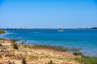 Portekiz, Alentejo 'daki Barragem de Morgavel rezervuarının manzarası, sakin mavi suyu, ön planda kayalık kıyı şeridini ve açık bir gökyüzünün altındaki ormanlık tepeleri gösteriyor..