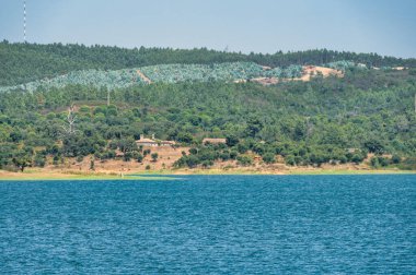 Portekiz, Alentejo 'daki Barragem de Morgavel rezervuarının manzarası, sakin mavi suyu, ön planda kayalık kıyı şeridini ve açık bir gökyüzünün altındaki ormanlık tepeleri gösteriyor..
