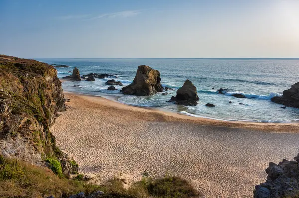 Portekiz 'in Alentejo kentindeki Praia da Samouqueira kumsalında büyük kaya oluşumlarına karşı okyanus dalgaları, parçalı bulutlu bir gökyüzünün altında engebeli bir kıyı manzarasını gösteriyor..