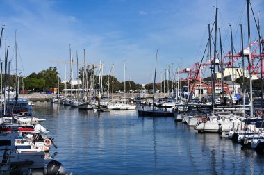 Açık güneşli bir günde limana demirlemiş birkaç yelkenli ve yat manzarası, arka planda vinçler ve deniz kıyısındaki binalar görülüyor..