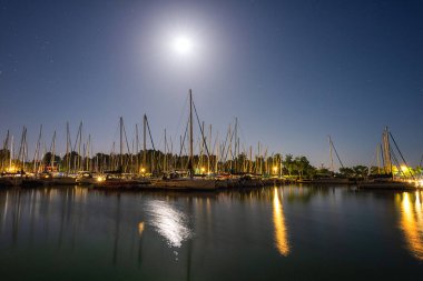 Macaristan 'daki Balaton Gölü, gece manzaralı. Yatlı manzara, deniz feneri, yıldızlı ve aylı koyu mavi gökyüzü, yansımalı sakin su, arka planda açık hava yolculuğu