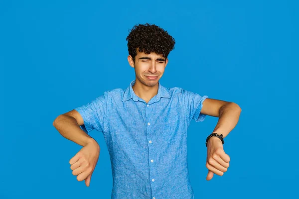 disappointed student showing thumbs down after watching his final exams, wearing casual clothes over blue isolated background