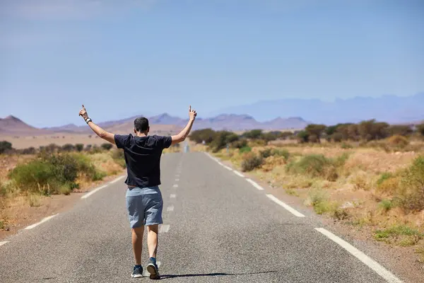 stock image A traveler raises his arms in victory while walking along an endless road through the Moroccan desert, symbolizing freedom and adventure.