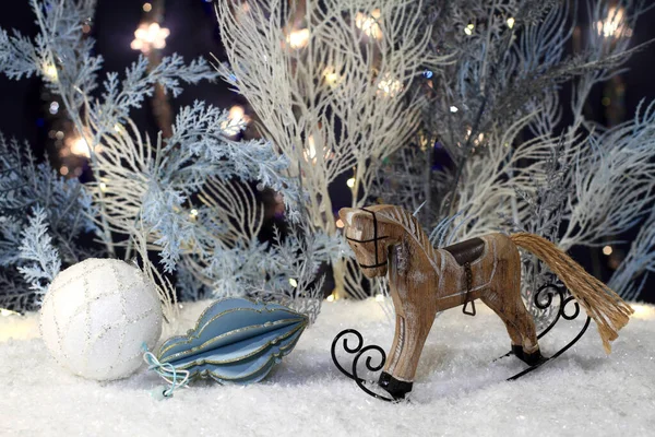 Wooden toy rocking horse stands on artificial snow and New Year toys lie next to it and artificial blue and white trees with festive lights stand behind them against a dark blue background. Closeup. Night studio shooting.