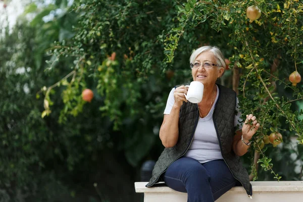 日当たりの良い庭でお茶を飲んでいるシニア灰色の髪の女性見事な田舎 隠遁と退職と幸せなシニアライフスタイルのコンセプト — ストック写真