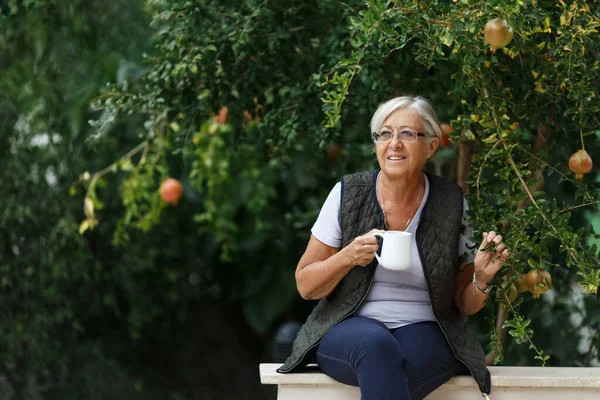 日当たりの良い庭でお茶を飲んでいるシニア灰色の髪の女性見事な田舎 隠遁と退職と幸せなシニアライフスタイルのコンセプト — ストック写真