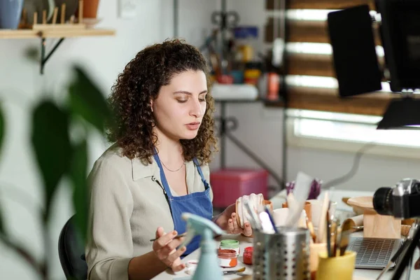 Females potter in blue apron making a vlog, recording online course, clay master class,lessons in her studion with earthenware shelf on background. Using led lamp,studio equipment.