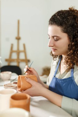 Concentrated woman painting clay jug with colourful paint shaping and sculpturing earthenware cup in pottery studio on ceramic workshop. Girl artist relaxing with art leisure.