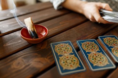 Mumun yanındaki masada tarot kartlarıyla falcı. Masaya sihirli otlar ve palo santo aroma çubukları serpiştirilmiş tarot kartları. Tahmin konsepti