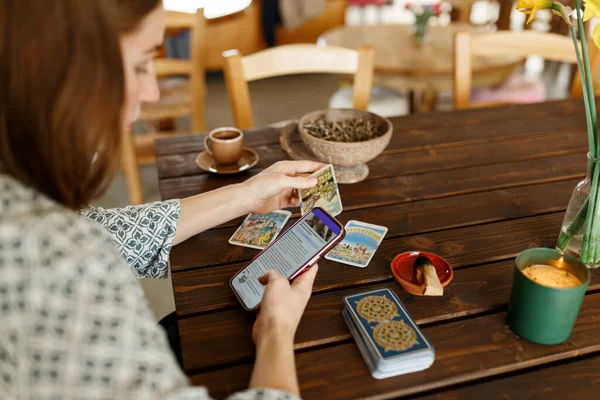 Güzel genç bir kadın, tarotlu kartlar üzerinde tahminde bulunuyor. Tahta masadaki rünler ve telefonda kehaneti, ev içini, mumları ve kuru otları yorumlamak için internet uygulaması kullanıyor...