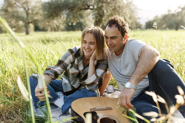 stock image Couple taking selfie or making video call while spending time together on picnic with guitar in green summer grass . Dating.