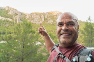 Mutlu yürüyüşçü bir selfie çeker ve parmağını dağlık araziyi gösterir.