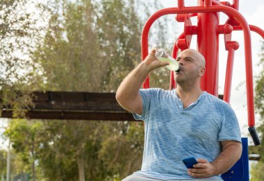Bir adam biyosağlık parkındaki makinede oturmuş izotonik spor içeceği içiyor.