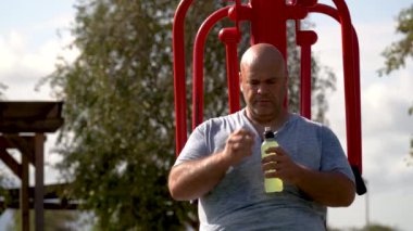 Bir adam spor suyu içiyor spor makinesinde rahatça oturuyor, açık havada..