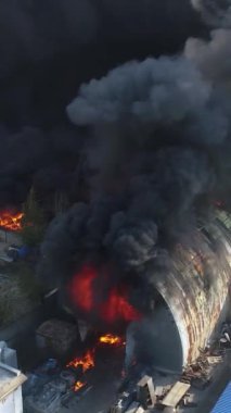 Büyük bir yangın deposunun insansız hava aracı videosu. Gökyüzünde yoğun siyah duman. Fabrika yanıyor. Depo endüstriyel hangarı. Kurtarma ekipleri yangını söndürüyor. Yukarıdan bak. Dikey video.