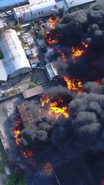 Büyük bir yangın deposunun insansız hava aracı videosu. Gökyüzünde yoğun siyah duman. Fabrika yanıyor. Depo endüstriyel hangarı. Kurtarma ekipleri yangını söndürüyor. Yukarıdan bak. Dikey video.