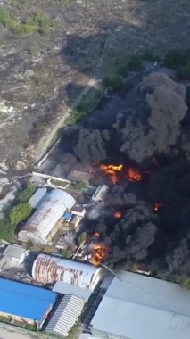 Büyük bir yangın deposunun insansız hava aracı videosu. Gökyüzünde yoğun siyah duman. Fabrika yanıyor. Depo endüstriyel hangarı. Kurtarma ekipleri yangını söndürüyor. Yukarıdan bak. Dikey video.