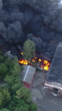 Büyük bir yangın deposunun insansız hava aracı videosu. Gökyüzünde yoğun siyah duman. Fabrika yanıyor. Depo endüstriyel hangarı. Kurtarma ekipleri yangını söndürüyor. Yukarıdan bak. Dikey video.