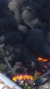 Büyük bir yangın deposunun insansız hava aracı videosu. Gökyüzünde yoğun siyah duman. Fabrika yanıyor. Depo endüstriyel hangarı. Kurtarma ekipleri yangını söndürüyor. Yukarıdan bak. Dikey video.