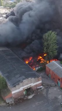 Büyük bir yangın deposunun insansız hava aracı videosu. Gökyüzünde yoğun siyah duman. Fabrika yanıyor. Depo endüstriyel hangarı. Kurtarma ekipleri yangını söndürüyor. Yukarıdan bak. Dikey video.