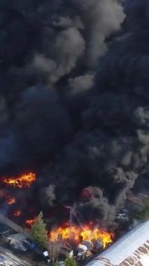 Büyük bir yangın deposunun insansız hava aracı videosu. Gökyüzünde yoğun siyah duman. Fabrika yanıyor. Depo endüstriyel hangarı. Kurtarma ekipleri yangını söndürüyor. Yukarıdan bak. Dikey video.
