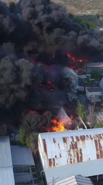 Büyük bir yangın deposunun insansız hava aracı videosu. Gökyüzünde yoğun siyah duman. Fabrika yanıyor. Depo endüstriyel hangarı. Kurtarma ekipleri yangını söndürüyor. Yukarıdan bak. Dikey video.