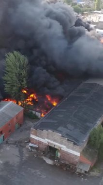 Büyük bir yangın deposunun insansız hava aracı videosu. Gökyüzünde yoğun siyah duman. Fabrika yanıyor. Depo endüstriyel hangarı. Kurtarma ekipleri yangını söndürüyor. Yukarıdan bak. Dikey video.