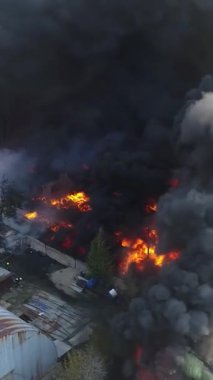 Büyük bir yangın deposunun insansız hava aracı videosu. Gökyüzünde yoğun siyah duman. Fabrika yanıyor. Depo endüstriyel hangarı. Kurtarma ekipleri yangını söndürüyor. Yukarıdan bak. Dikey video.