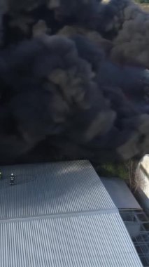 Büyük bir yangın deposunun insansız hava aracı videosu. Gökyüzünde yoğun siyah duman. Fabrika yanıyor. Depo endüstriyel hangarı. Kurtarma ekipleri yangını söndürüyor. Yukarıdan bak. Dikey video.