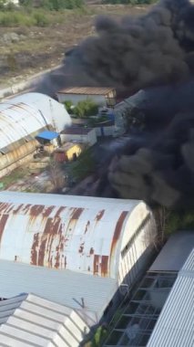Büyük bir yangın deposunun insansız hava aracı videosu. Gökyüzünde yoğun siyah duman. Fabrika yanıyor. Depo endüstriyel hangarı. Kurtarma ekipleri yangını söndürüyor. Yukarıdan bak. Dikey video.