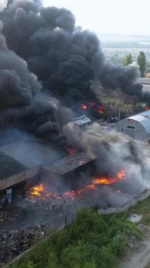 Büyük bir yangın deposunun insansız hava aracı videosu. Gökyüzünde yoğun siyah duman. Fabrika yanıyor. Depo endüstriyel hangarı. Kurtarma ekipleri yangını söndürüyor. Yukarıdan bak. Dikey video.