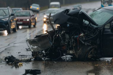 Islak bir yolda kaza yaptıktan sonra kaza yapmış siyah bir araba. Ön tarafı parçalanmış ve kaputu açık bir araba şehir caddesinde. Yağmurlu bir hava. İki araba arasındaki çarpışmadan sonra...
