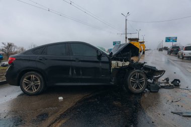 Islak bir yolda kaza yaptıktan sonra kaza yapmış siyah bir araba. Ön tarafı parçalanmış ve kaputu açık bir araba şehir caddesinde. Yağmurlu bir hava. İki araba arasındaki çarpışmadan sonra...