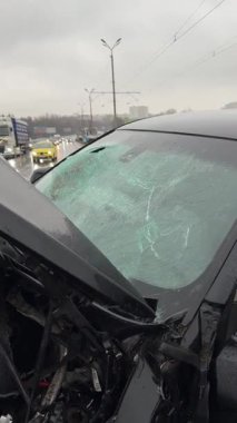 Islak bir yolda kaza yaptıktan sonra kaza yapmış siyah bir araba. Ön tarafı parçalanmış ve kaputu açık bir araba şehir caddesinde. Yağmurlu bir hava. İki araba arasındaki çarpışmadan sonra...