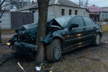 Ön tarafı ezilmiş ve kaputu açık siyah bir araba bir şehir caddesinde. Yakınlarda arabanın çarptığı bir ağaç var. Kazadan sonra..