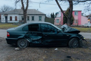 Ön tarafı ezilmiş ve kaputu açık siyah bir araba bir şehir caddesinde. Yakınlarda arabanın çarptığı bir ağaç var. Kazadan sonra..