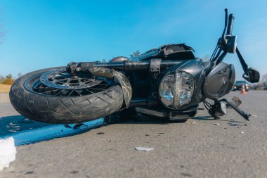 Asfaltta ciddi hasar görmüş siyah bir motosiklet var. Kazaya yakın çekim. Kontrolü kaybettim ve düştüm. Araba sürme tehlikesi. Başka bir araca çarpmış. Kırık farlar ve eğilmiş gidonlar..
