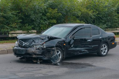 Çarpışmadan sonra ön tarafı kırılmış siyah bir sedan. Tamponda, kaputta ve farlarda hasar var. Asfaltta kırıntılar ve sıvı izleri var.