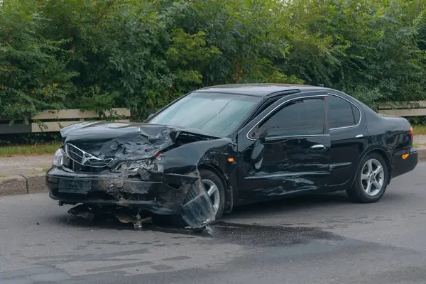 Çarpışmadan sonra ön tarafı kırılmış siyah bir sedan. Tamponda, kaputta ve farlarda hasar var. Asfaltta kırıntılar ve sıvı izleri var.