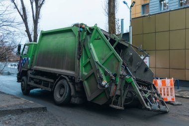 Bir çukura düştükten sonra yeşil bir çöp kamyonu bir şehir caddesinde yan yatar. Turuncu uyarı ağı asfalt yolda bir delik olduğunu gösteriyor. Yolda bir çukur